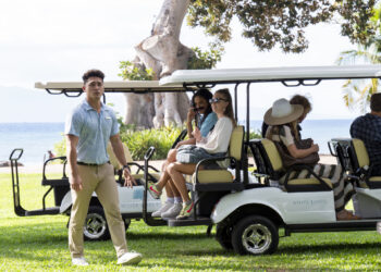 Kekoa Kekumano, Brittany O'Grady and Sydney Sweeney on the set of The White Lotus season 1. The shot shows them on the bag of a golf cart just after arriving at the hotel