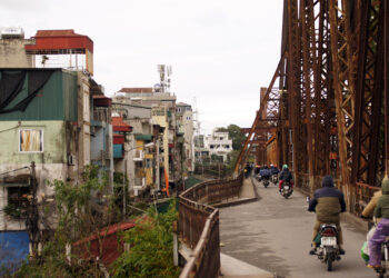 View of Long Biên Bridge. Half the image shows scooters passing along the bridge, while the other half shows nearby houses